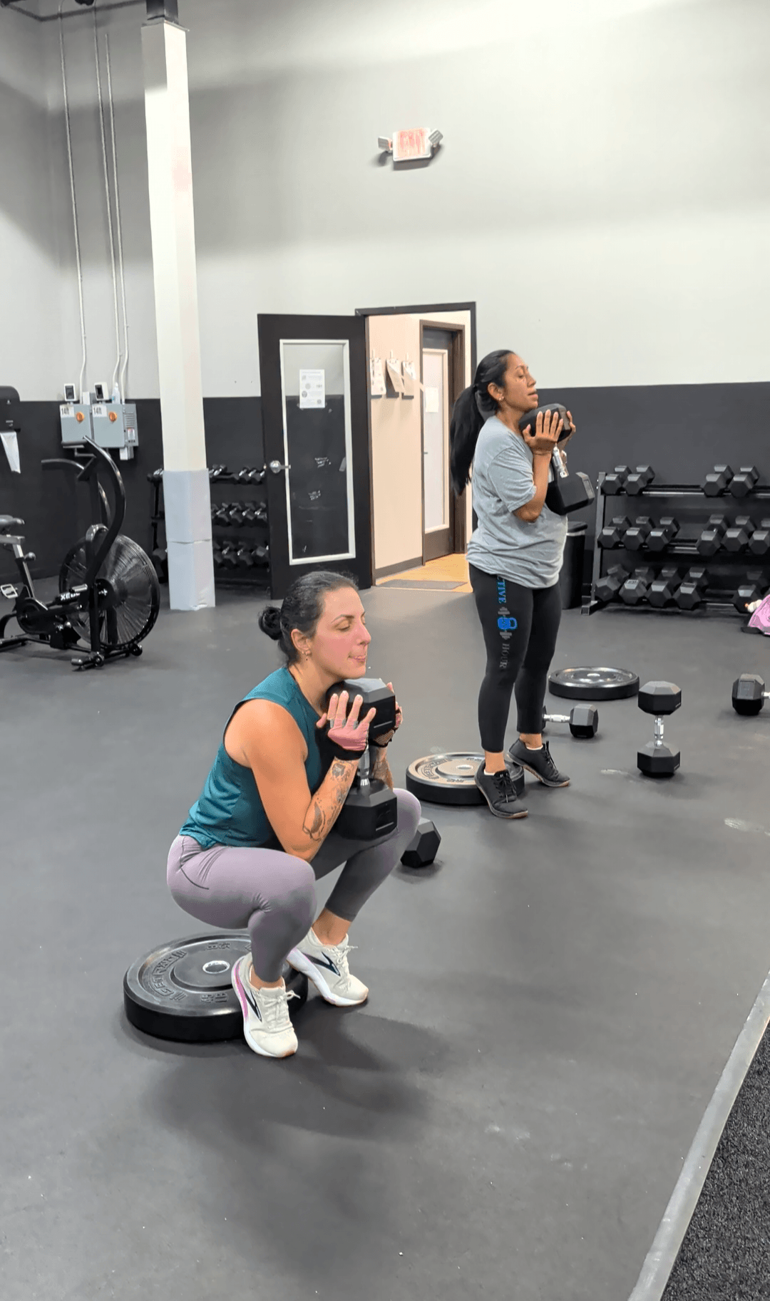Two women in a gym, one squatting with a weight plate, the other standing and drinking water, gym equipment visible.