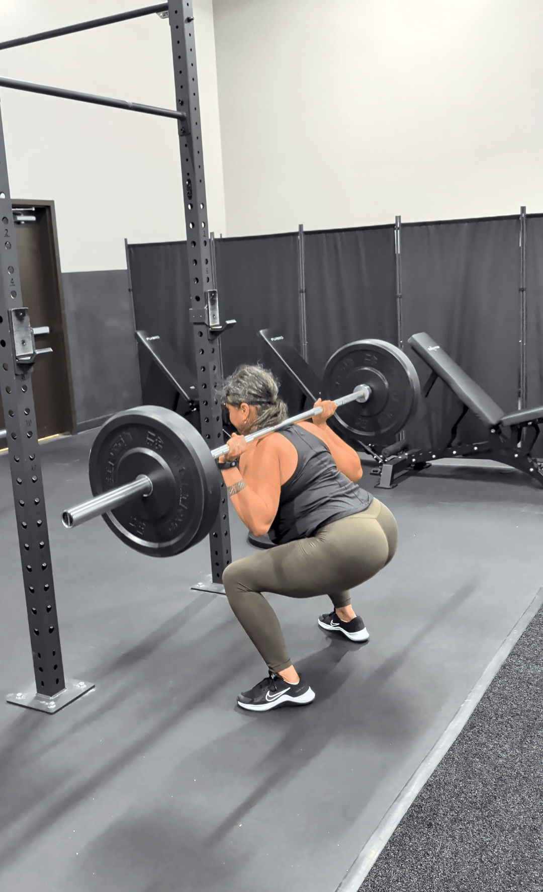 Person performing a squat exercise with a barbell in a gym, facing sideways, with gym equipment in background.