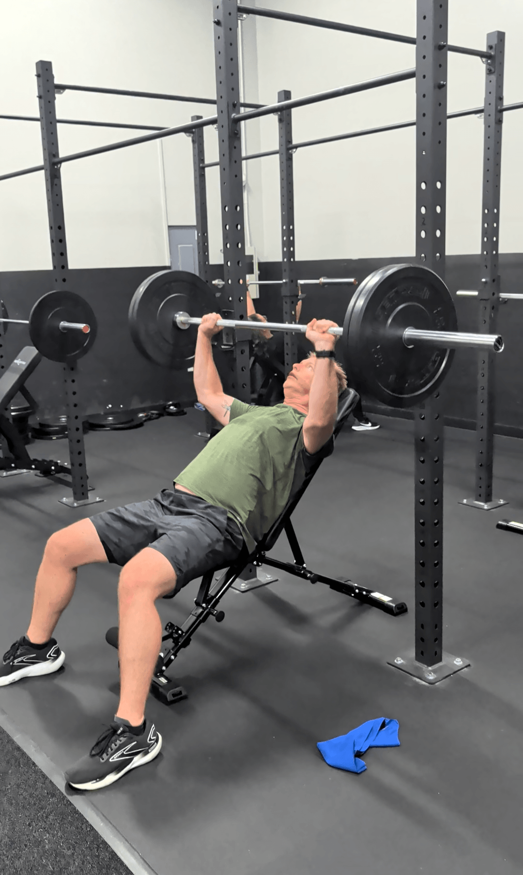 Person lying on bench pressing a barbell with weights in a gym, with equipment and racks in background.