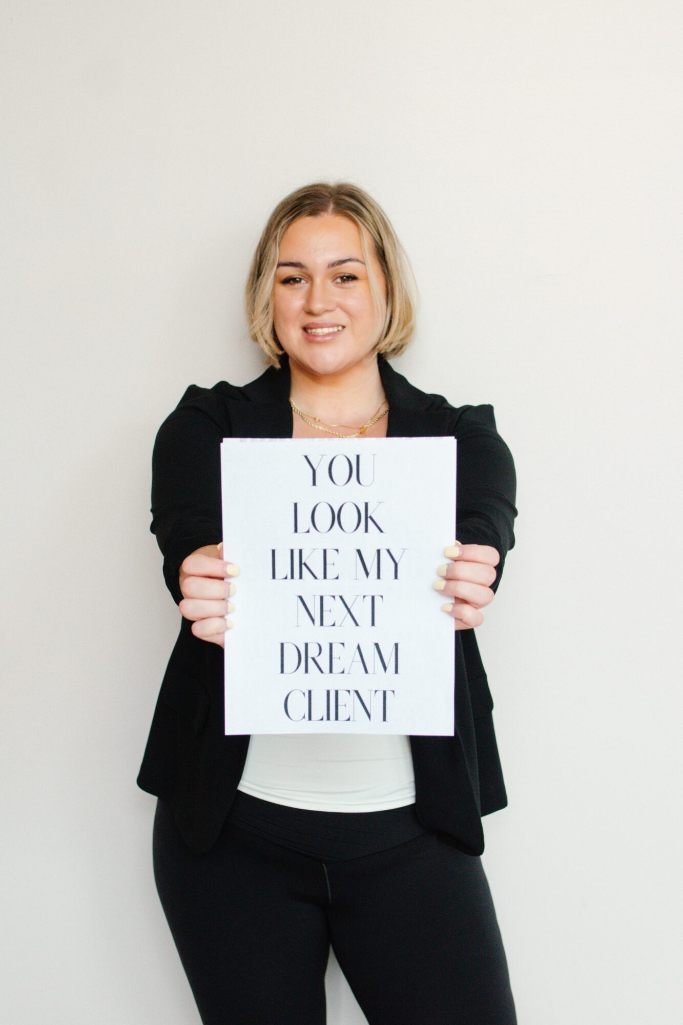 Young woman with blonde hair holding a sign that reads, 'You look like my next dream client,' smiling, standing against a plain white wall.