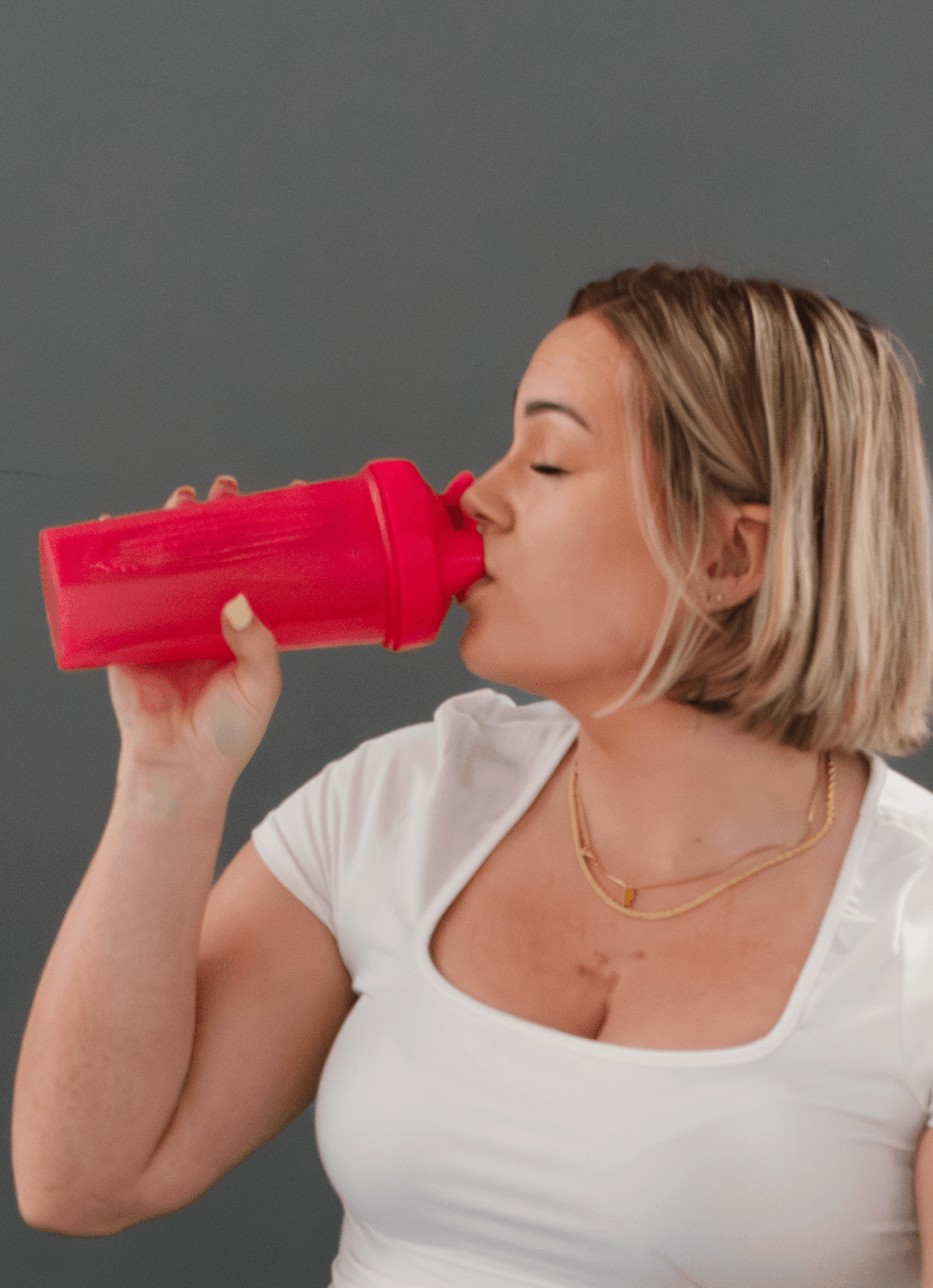Woman with short blonde hair drinking from a red water bottle, wearing a white top and gold necklace.