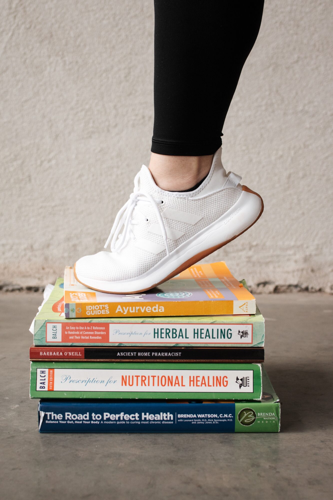 Person standing on a stack of health and wellness books, wearing white sneakers and black leggings, against a plain wall.