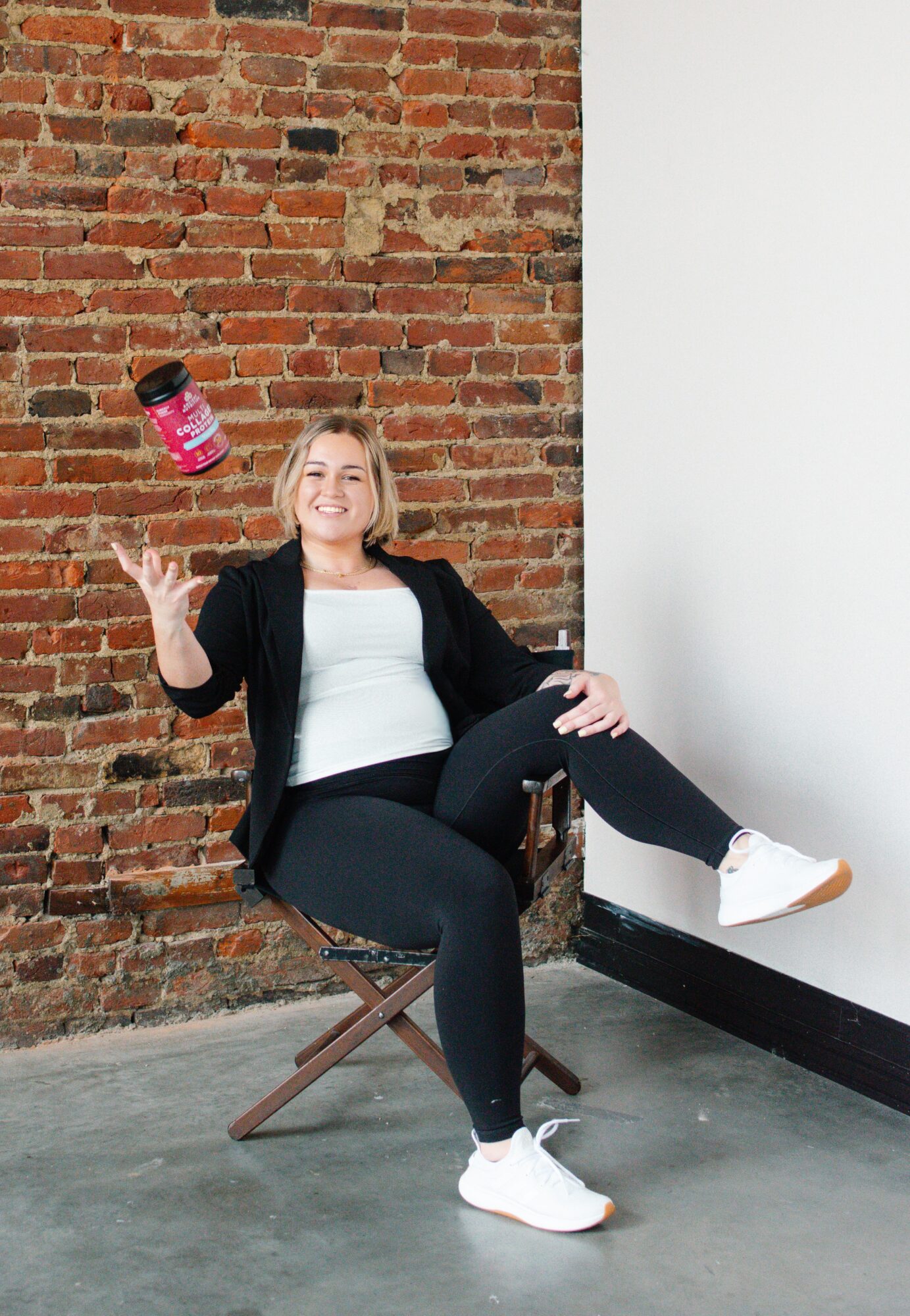 Woman sitting on a wooden chair, tossing a small jar in the air, smiling, against a brick wall and white wall background.