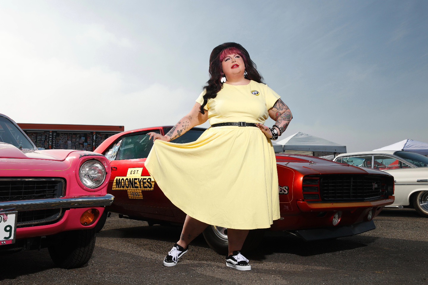 Person with long dark hair in a yellow dress standing between vintage cars in a parking lot under a cloudy sky.