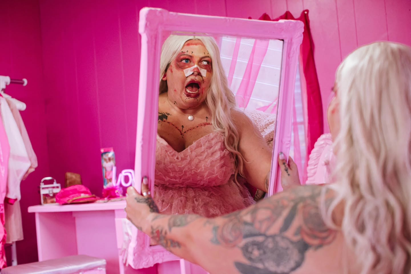 Person with long blonde hair and tattoos looking into a pink mirror, making a surprised face, in a pink room.