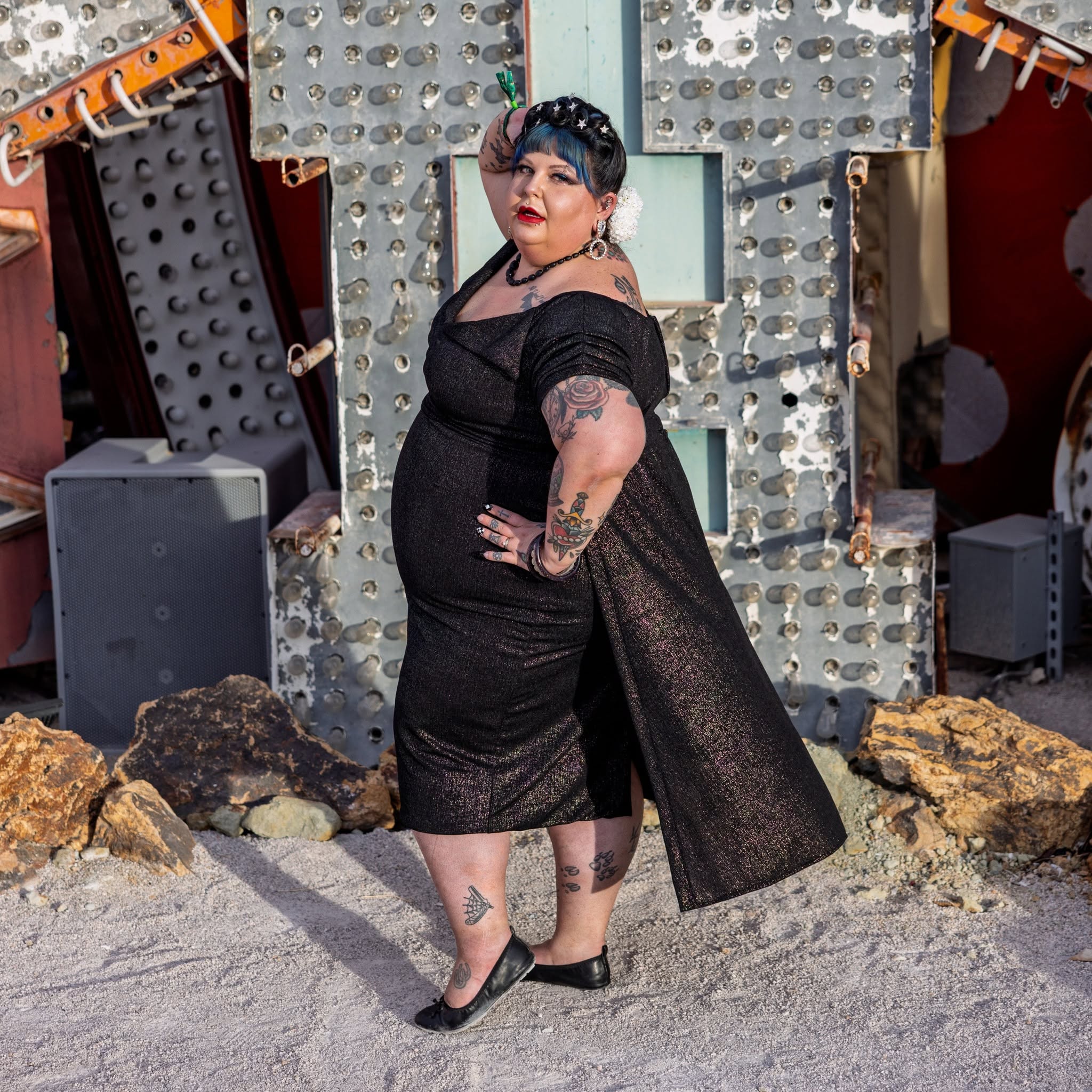 Person with dark hair and tattoos in a black dress and heels, standing outdoors with rocks and decorative background.