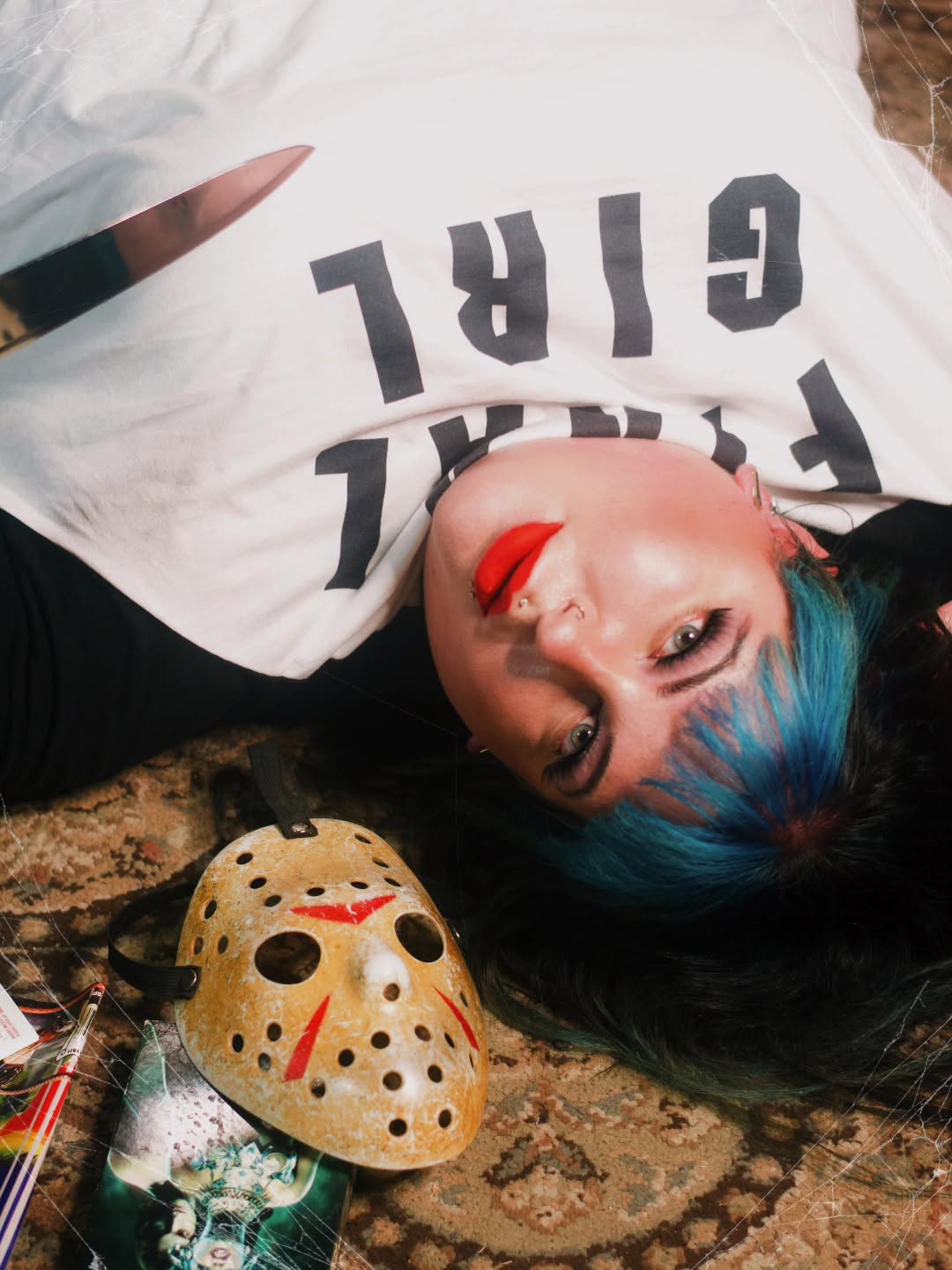 Person with blue hair lying on a patterned surface, wearing a white shirt with black text, next to a hockey mask and makeup tools.