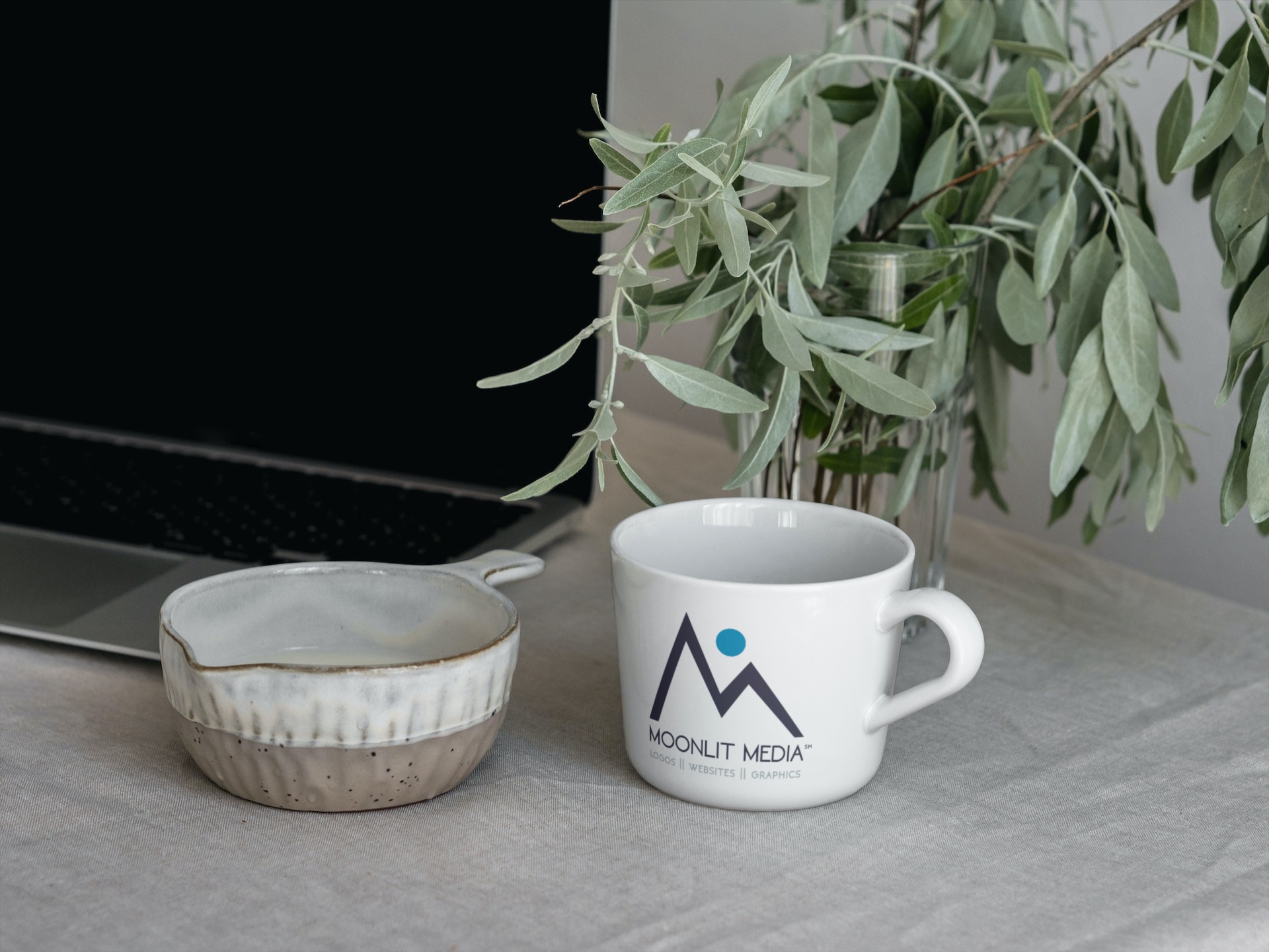 Coffee mug with logo, ceramic bowl, and plant on a table, with a laptop in the background.
