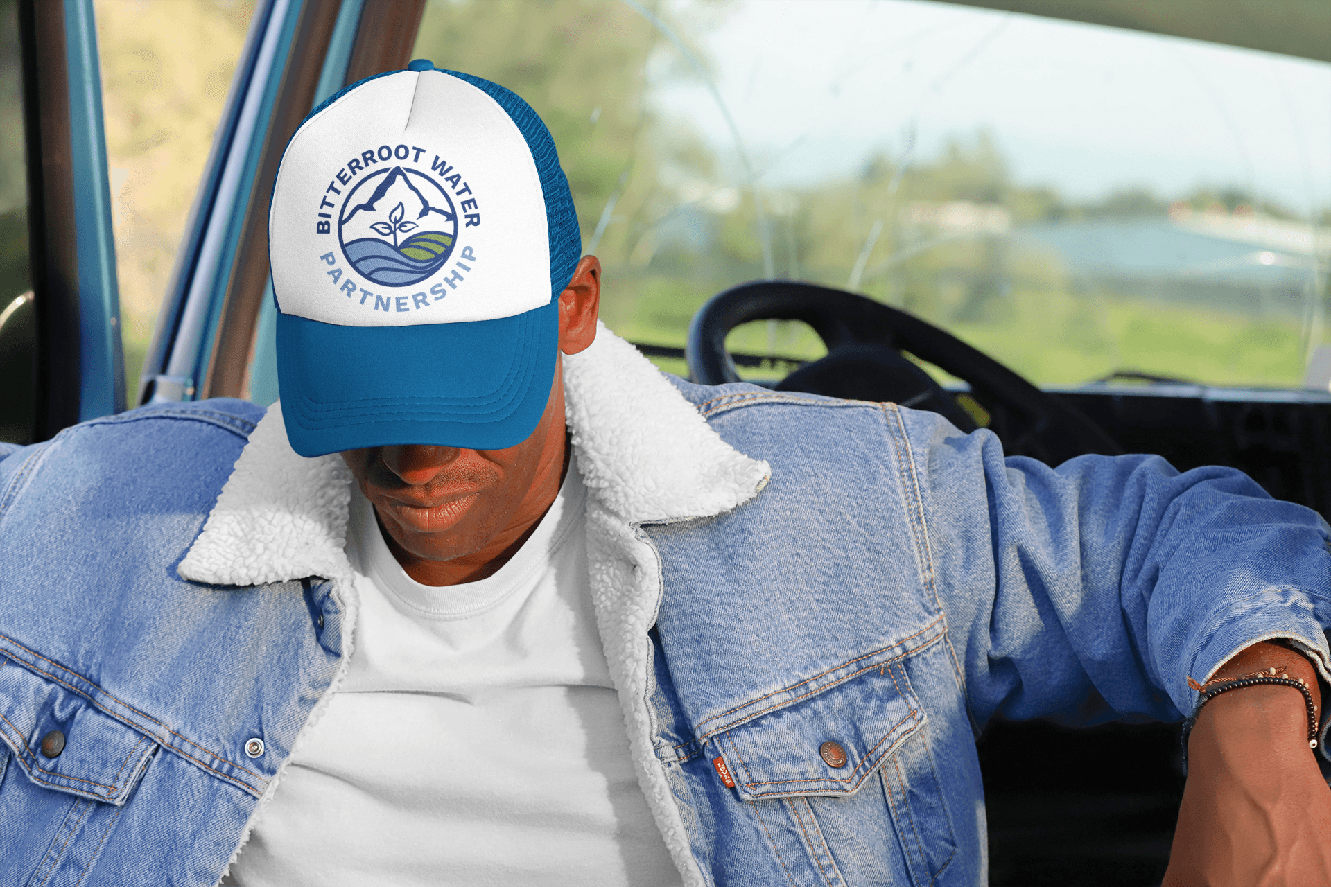 Person wearing a blue and white cap and denim jacket sitting in a vehicle, with a blurred outdoor background.