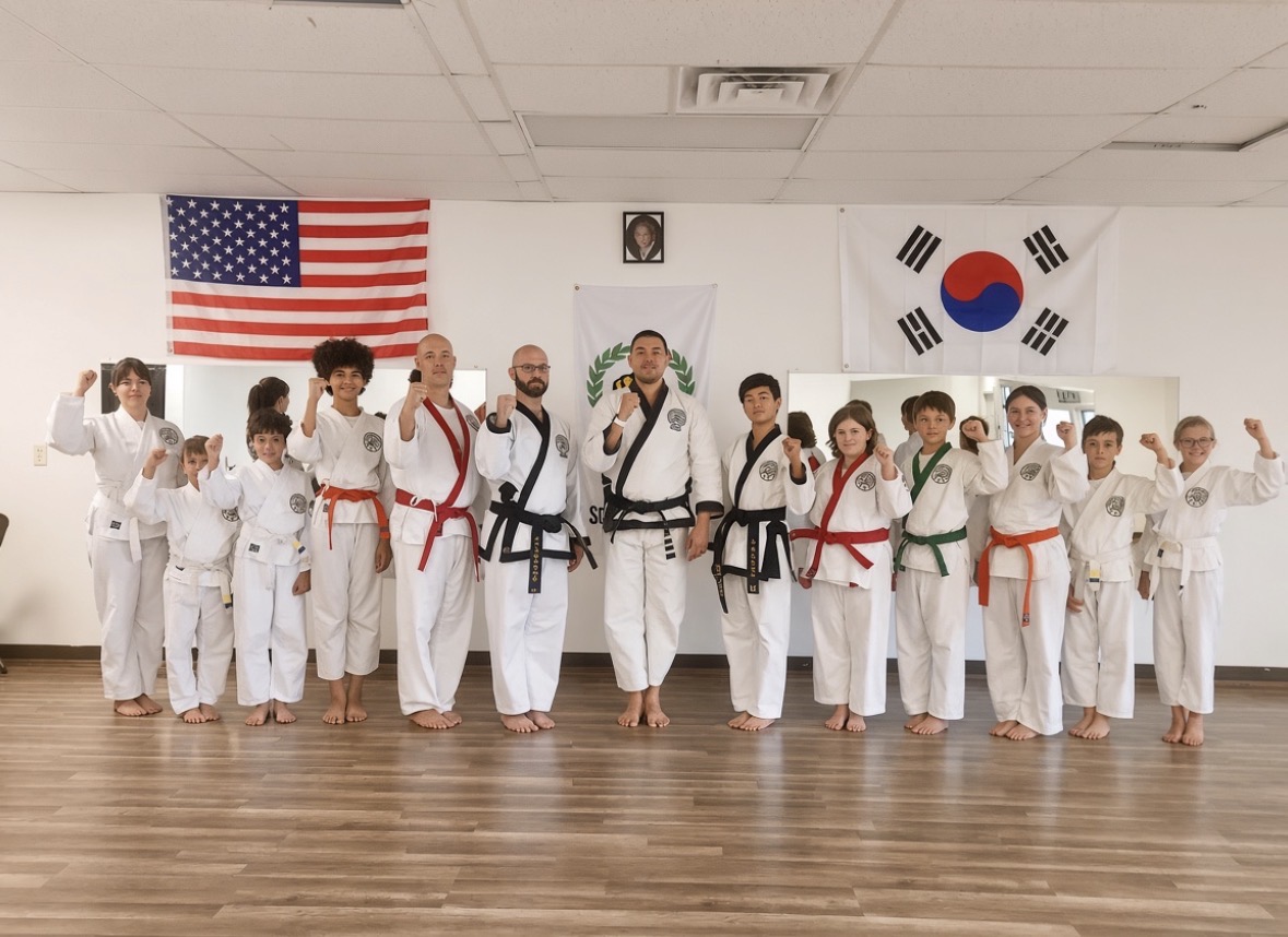 Group of children and instructors in martial arts uniforms standing in a dojo, flags of the United States and South Korea on the wall.