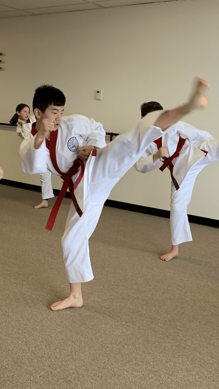 Group of children practicing martial arts in white uniforms with red belts, performing high kicks in a room with plain walls.