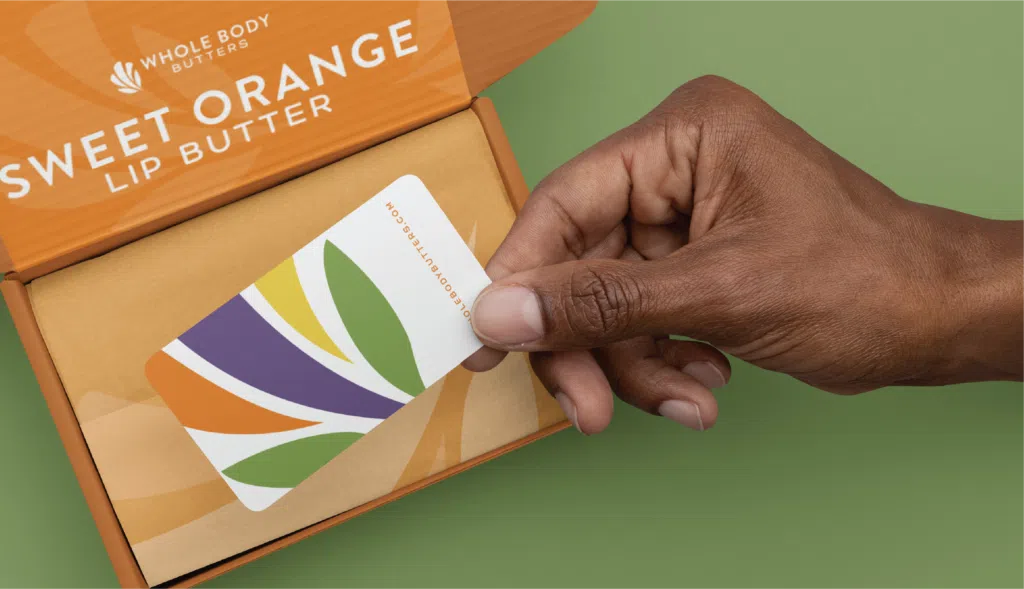 Hand holding a colorful card with leaf designs over an orange box labeled 'Sweet Orange Lip Butter'.