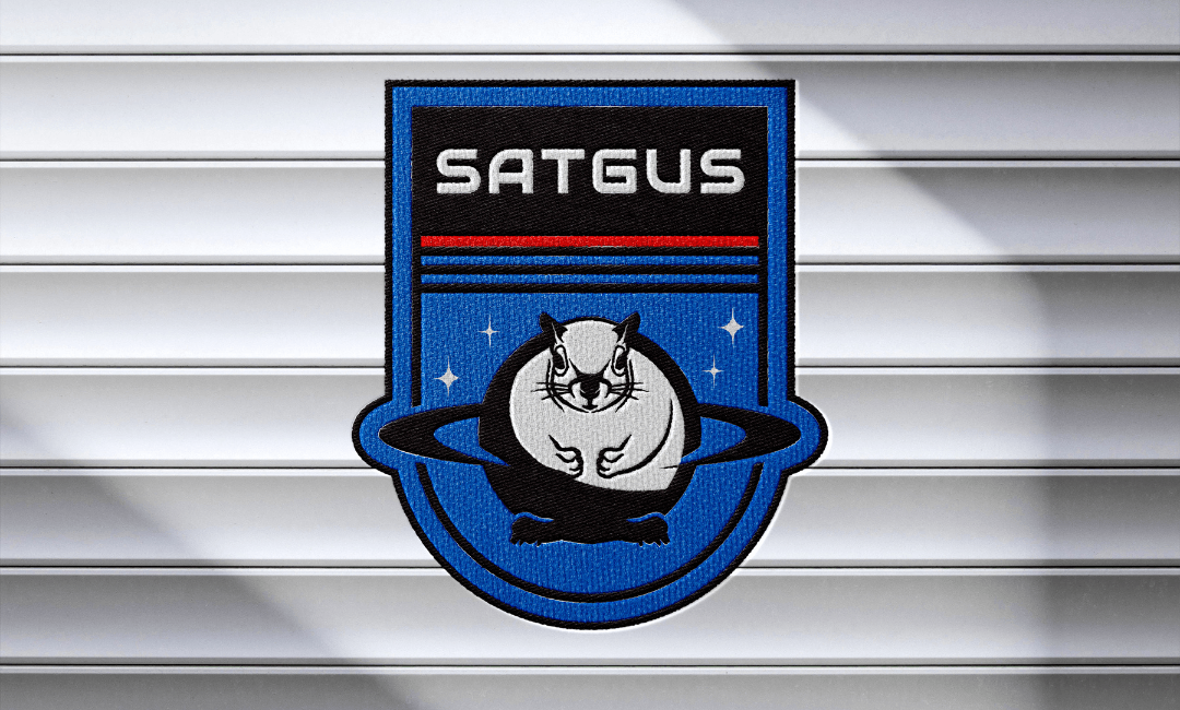 Patch with a cat wearing glasses, surrounded by a blue ring, and the word SATGUS at the top.