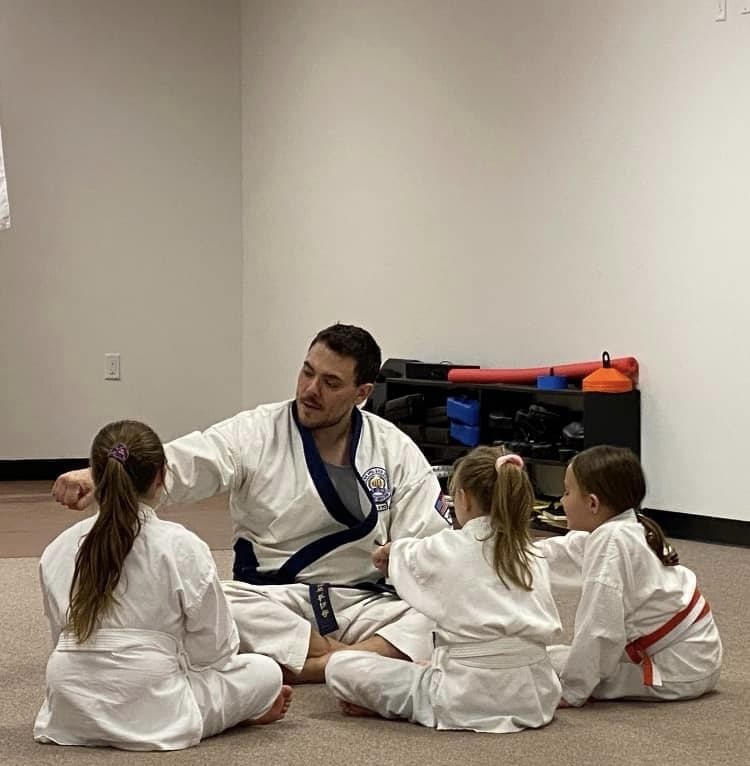 Instructor teaching children in martial arts class, sitting on the floor in a room with white walls and martial arts equipment.