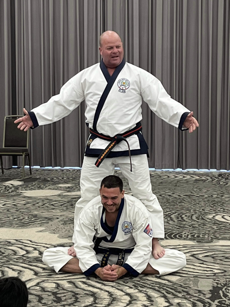 Two martial artists in gi uniforms, one standing with arms outstretched, the other kneeling in front, smiling, on a patterned carpet with curtains behind.