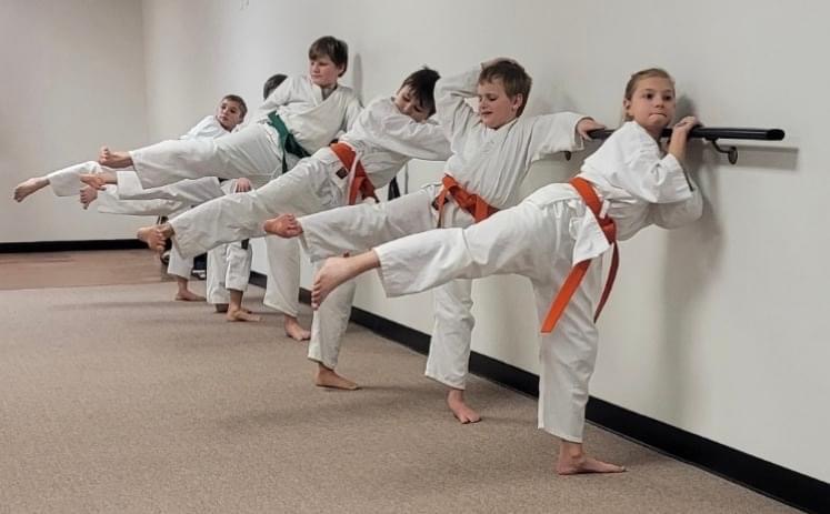 Six children in karate uniforms practicing kicks against a wall in a dojo, with one child holding a black stick.