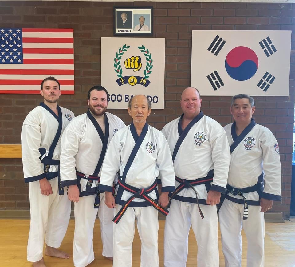 Five martial artists in uniforms stand in front of flags and emblems on a brick wall.