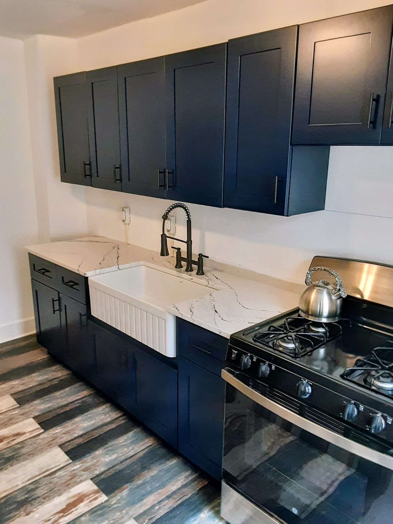 Kitchen with dark blue cabinets, white marble countertop, white farmhouse sink, and stainless steel stove, with wood-patterned flooring.