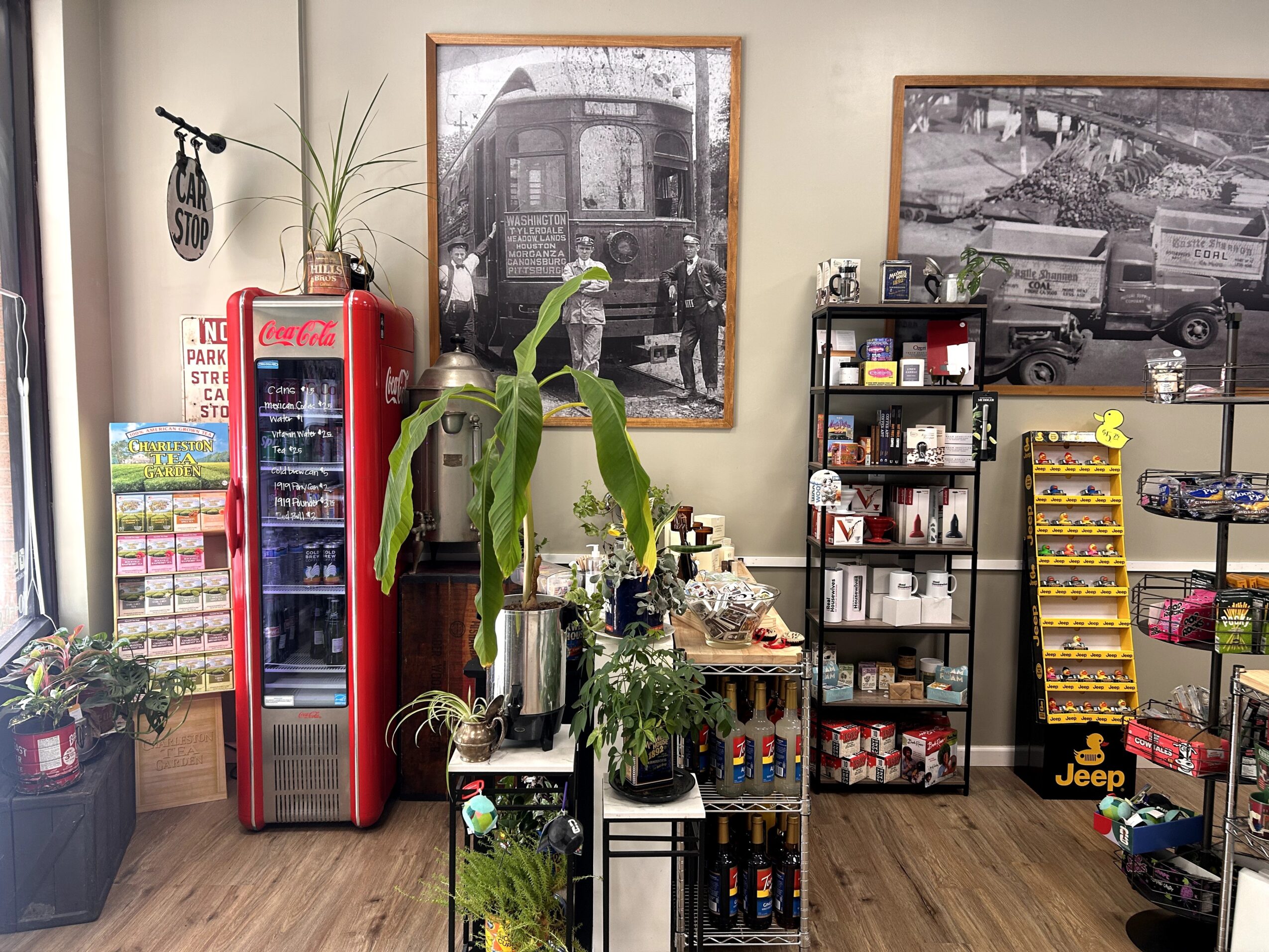 Indoor scene with a Coca-Cola refrigerator, plants, black and white photos, shelves with snacks and car accessories, wooden floor.