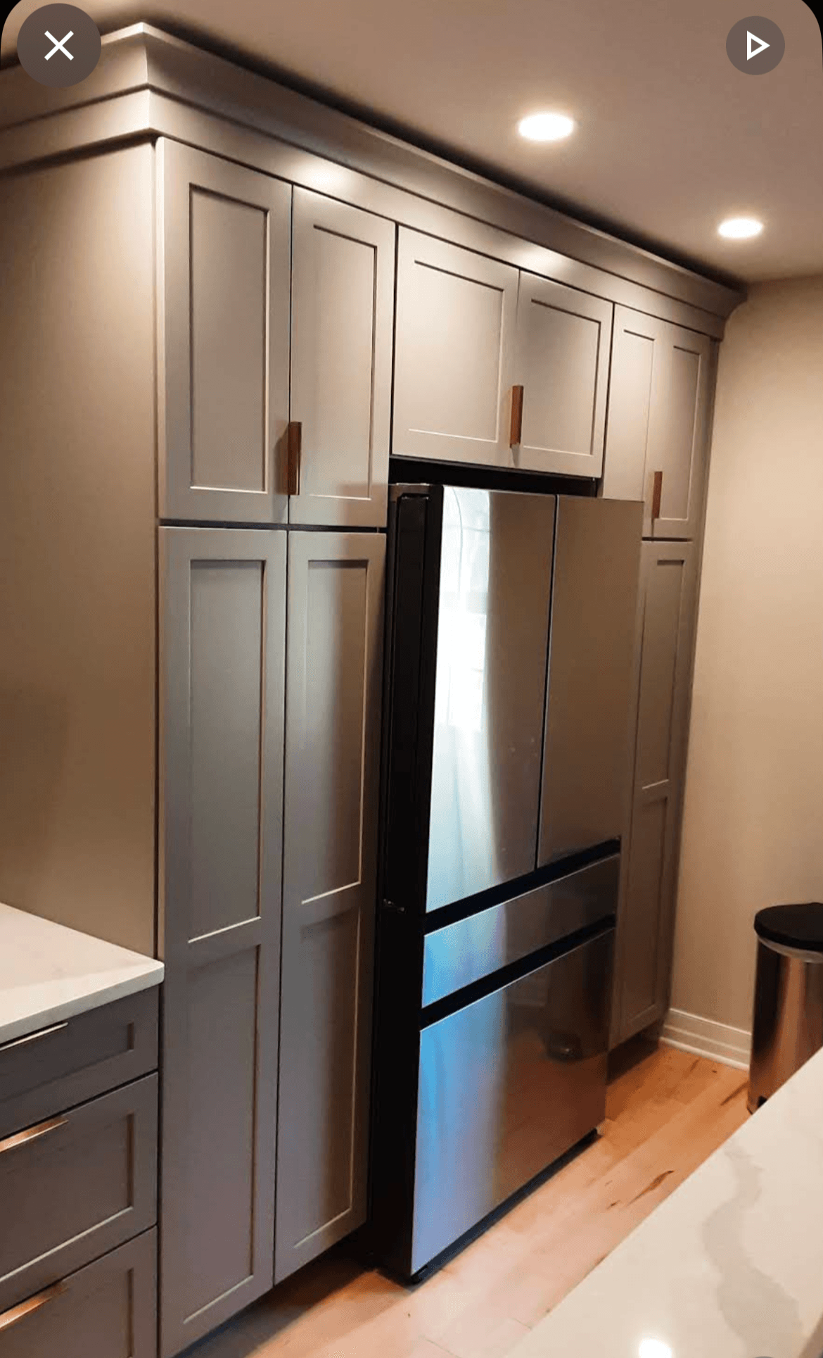 Tall gray kitchen cabinets with a stainless steel refrigerator in the center, in a modern kitchen with ceiling lights.