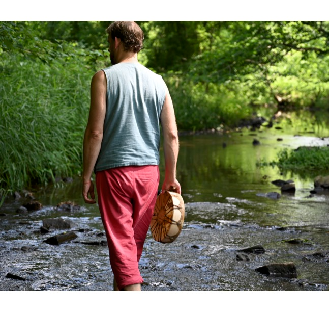 Person walking through a shallow stream holding a hat, surrounded by green trees and foliage.