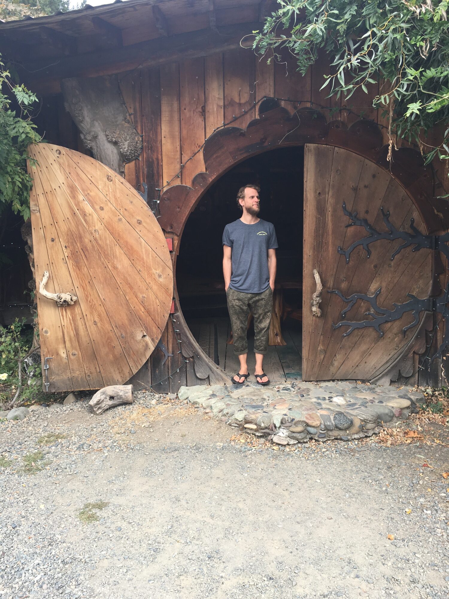 Man standing in front of a wooden circular door with animal carvings, surrounded by greenery and a rustic setting.
