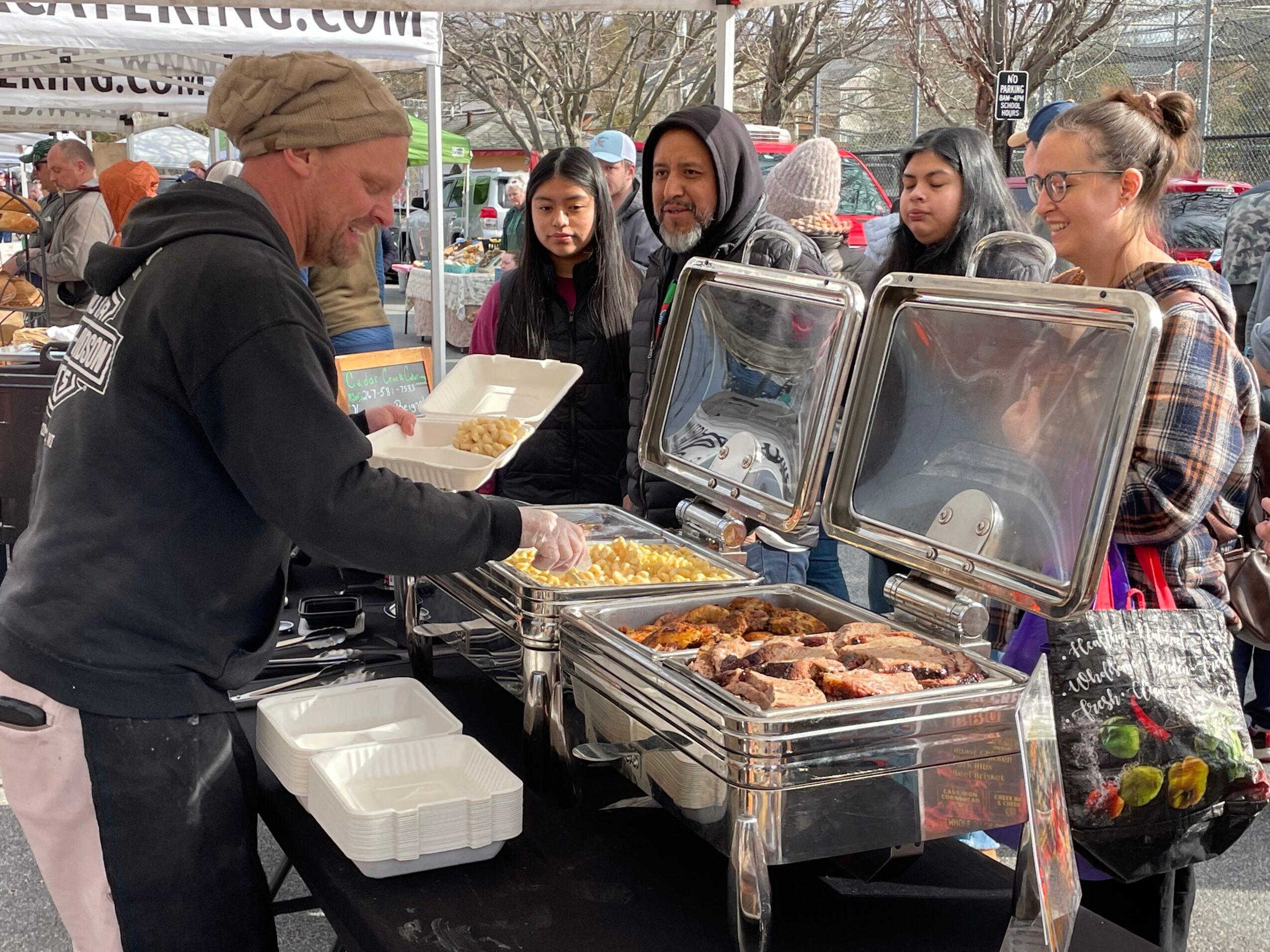 A woman serving food at a buffet-style outdoor event with four women waiting in line, food trays, and a tent in the background.