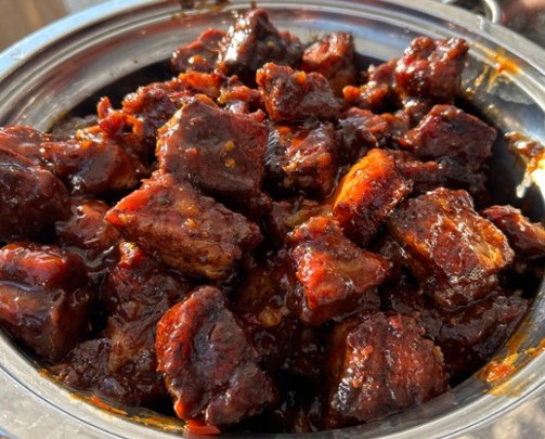 Close-up of cooked meat chunks in a metal dish, coated in dark sauce, with some orange-colored pieces visible.