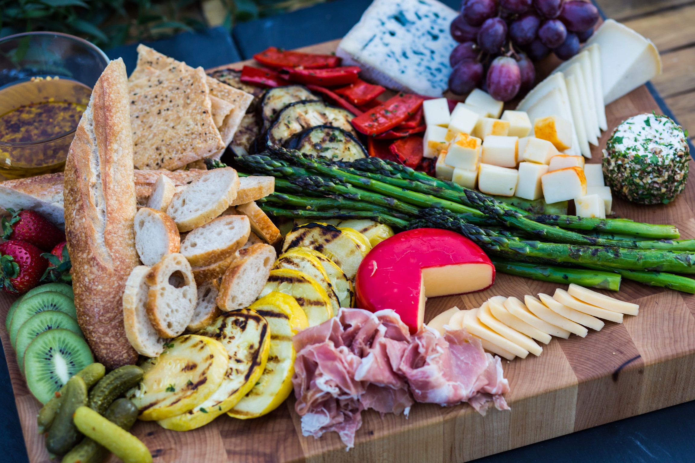 Assorted cheeses, grapes, bread, sliced meats, vegetables, and fruits on a wooden board.