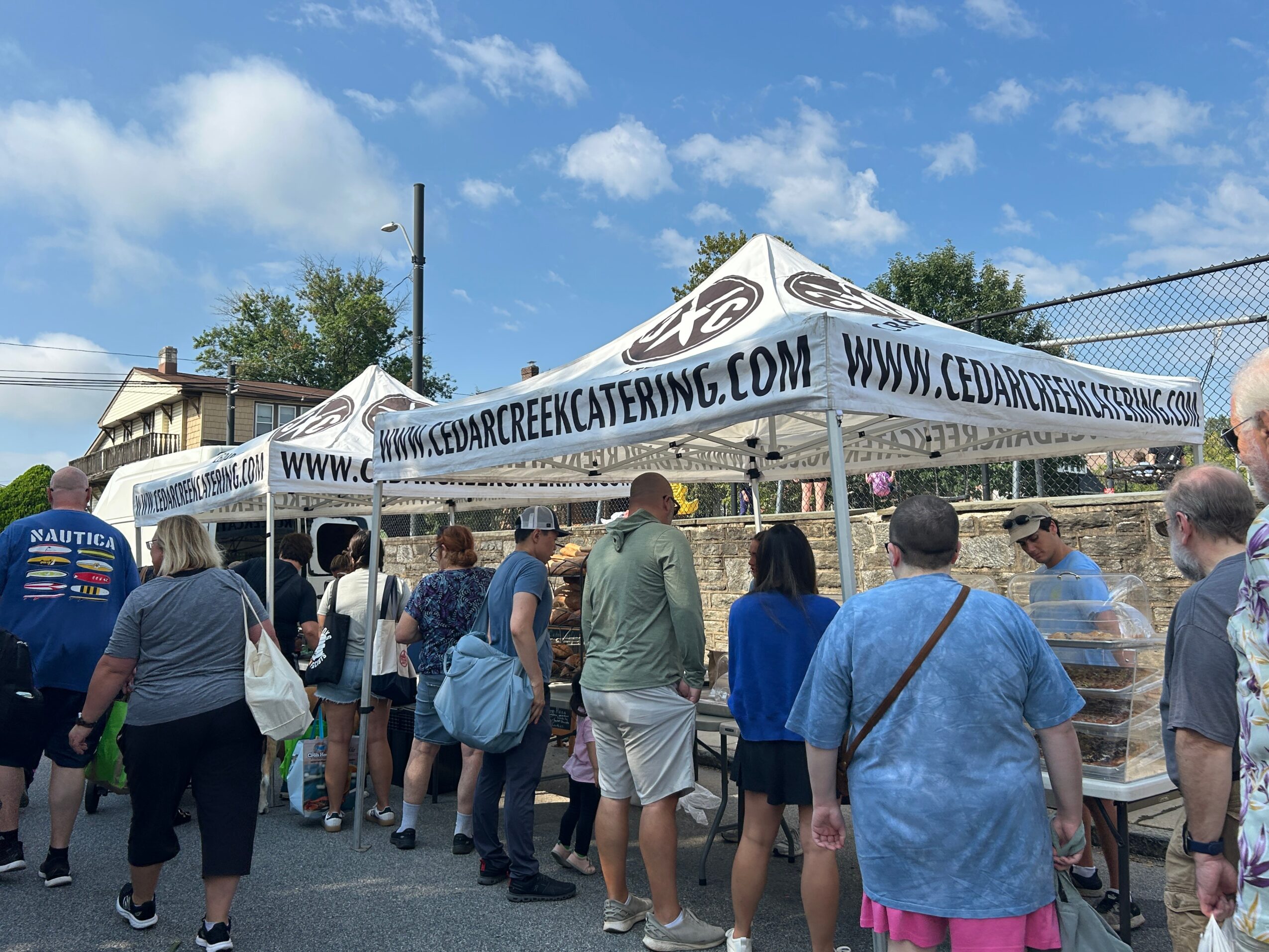 People line up at outdoor food tents with white canopies, blue sky, and trees in background.