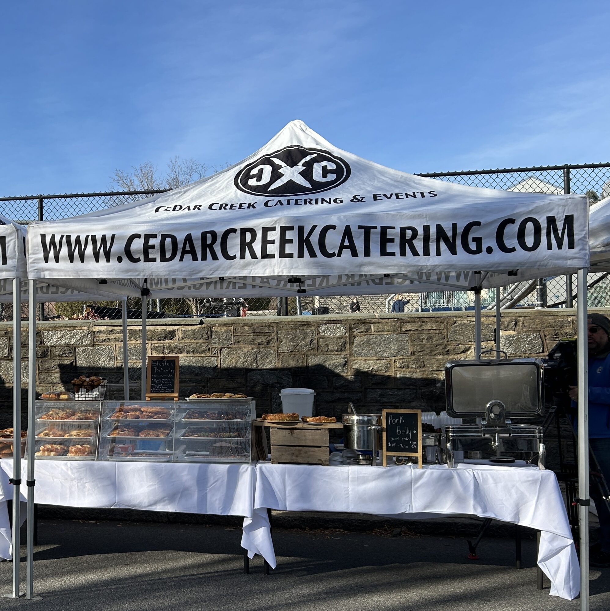 Outdoor catering booth with a white tent and table, food containers, and a banner displaying website and logo.