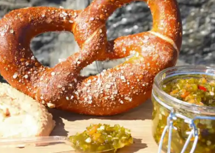 A large pretzel sprinkled with coarse salt, with a jar of relish and a bread roll nearby, outdoors.
