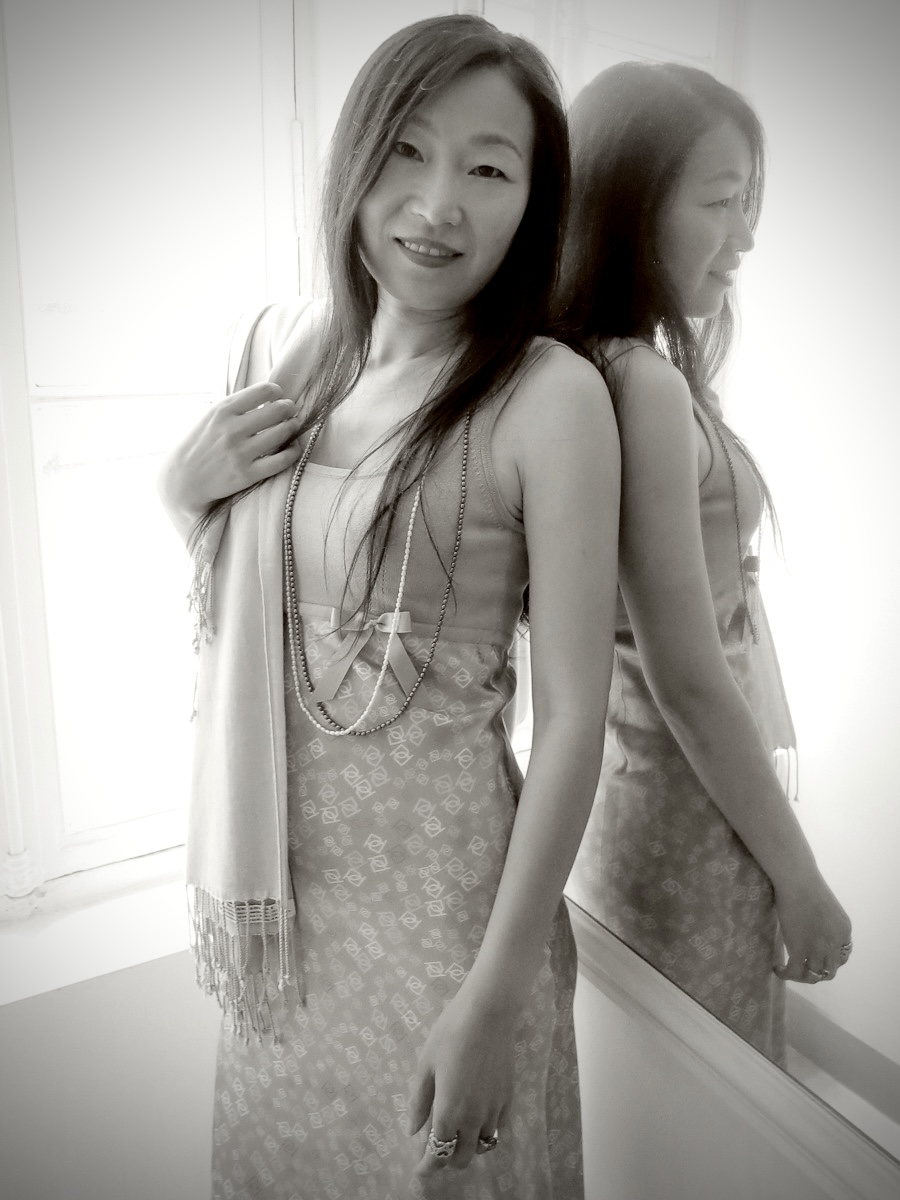 A woman stands in front of a mirror, smiling, with her reflection visible. She wears a dress and has long hair.