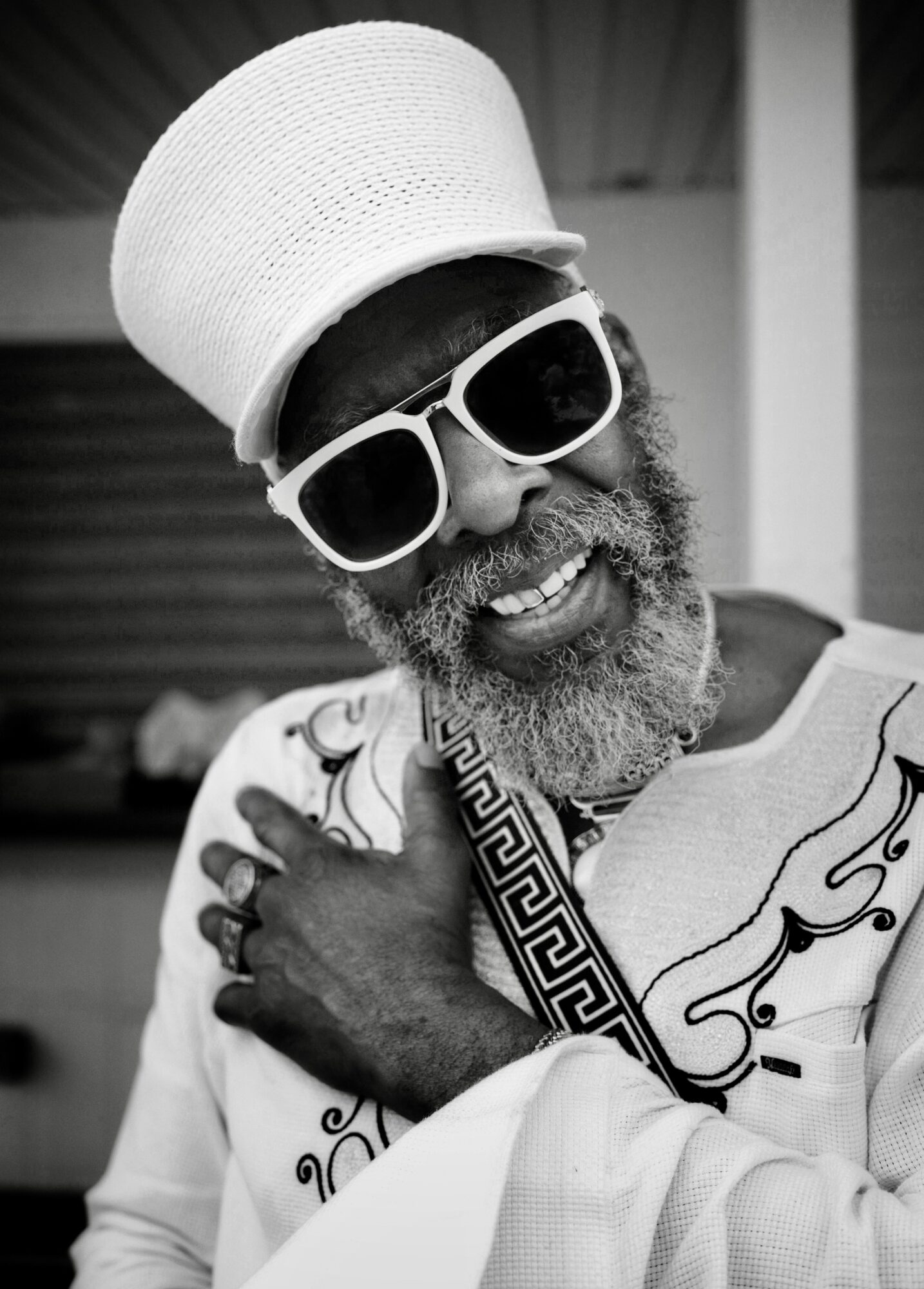 Person with large white hat, sunglasses, and patterned clothing, smiling and holding their shoulder.