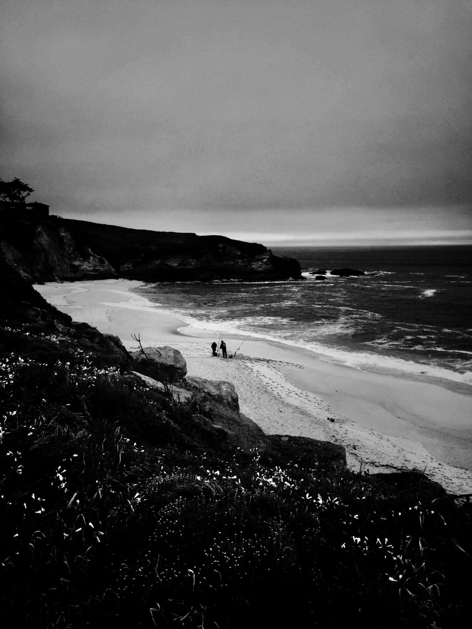 Coastal scene with cliffs, beach, and ocean, three people walking on the shore, cloudy sky, black and white image.