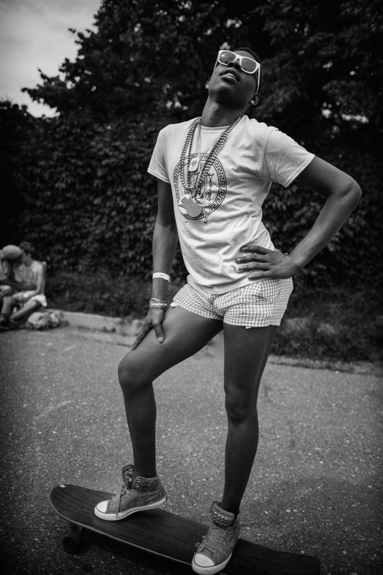 Person standing on skateboard outdoors, wearing glasses, t-shirt, shorts, and sneakers, with hand on hip and trees in background.