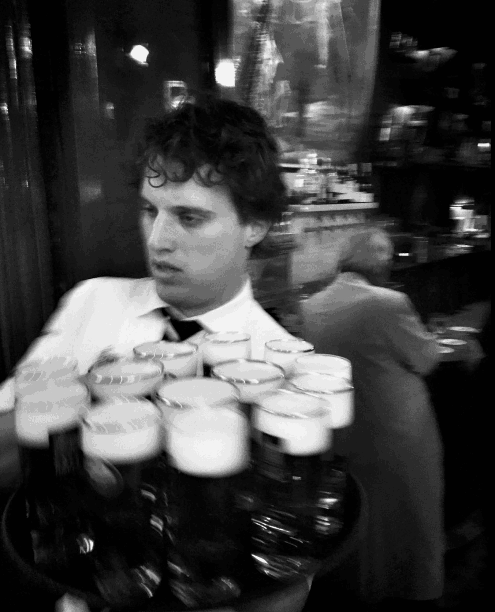 Young man with curly hair in a white shirt holding a tray of drinks in a bar or restaurant setting.