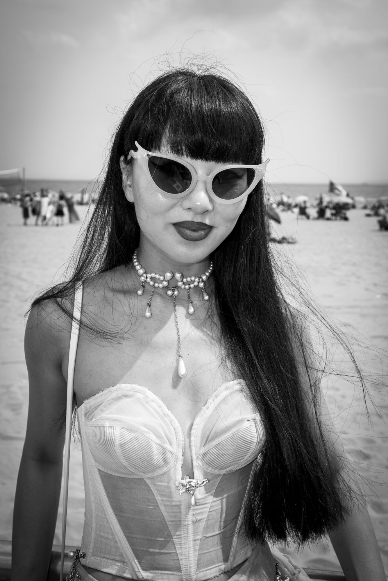 Woman with long hair, wearing sunglasses, a pearl necklace, and a strapless top at the beach.