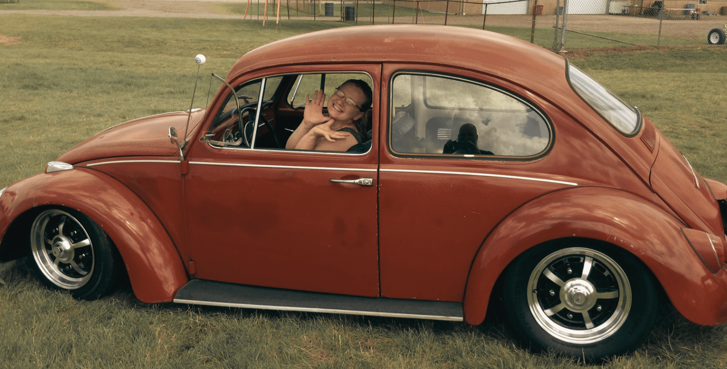 Child sitting inside a vintage brown car parked on grass, with a fence and field in the background.