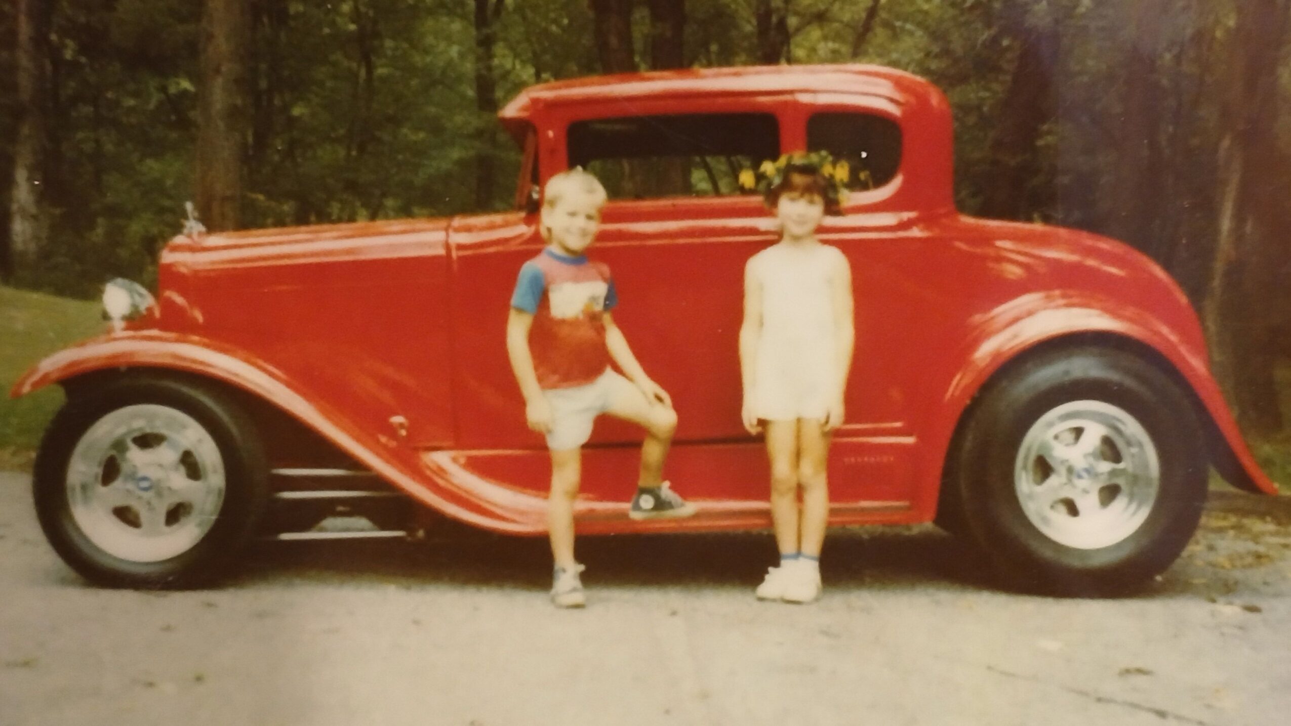 Two children sit on a red toy car outdoors, with trees in the background, upside down in the image.