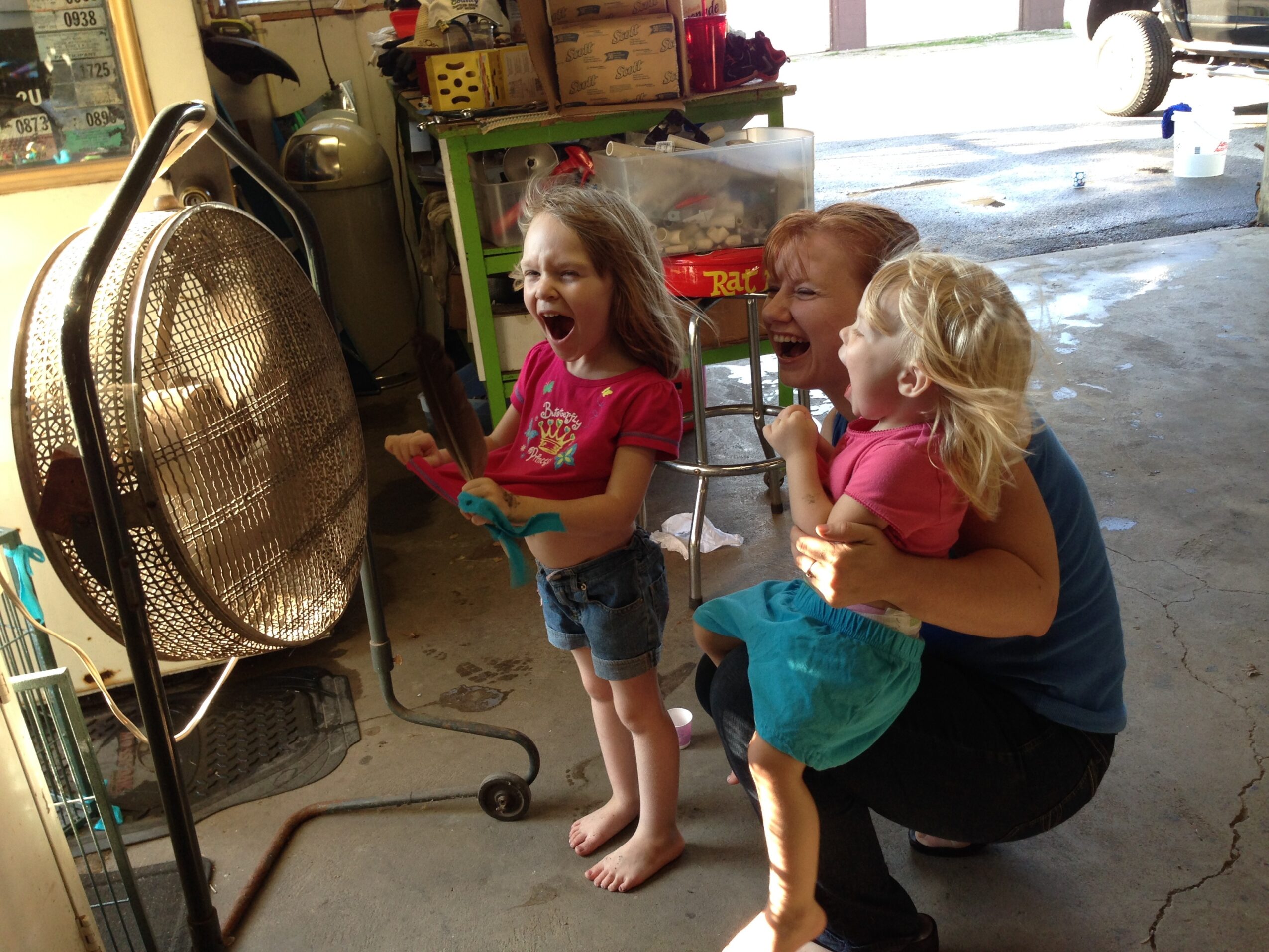 Three children watching a fan in a garage, smiling and laughing, with shelves and boxes in the background.