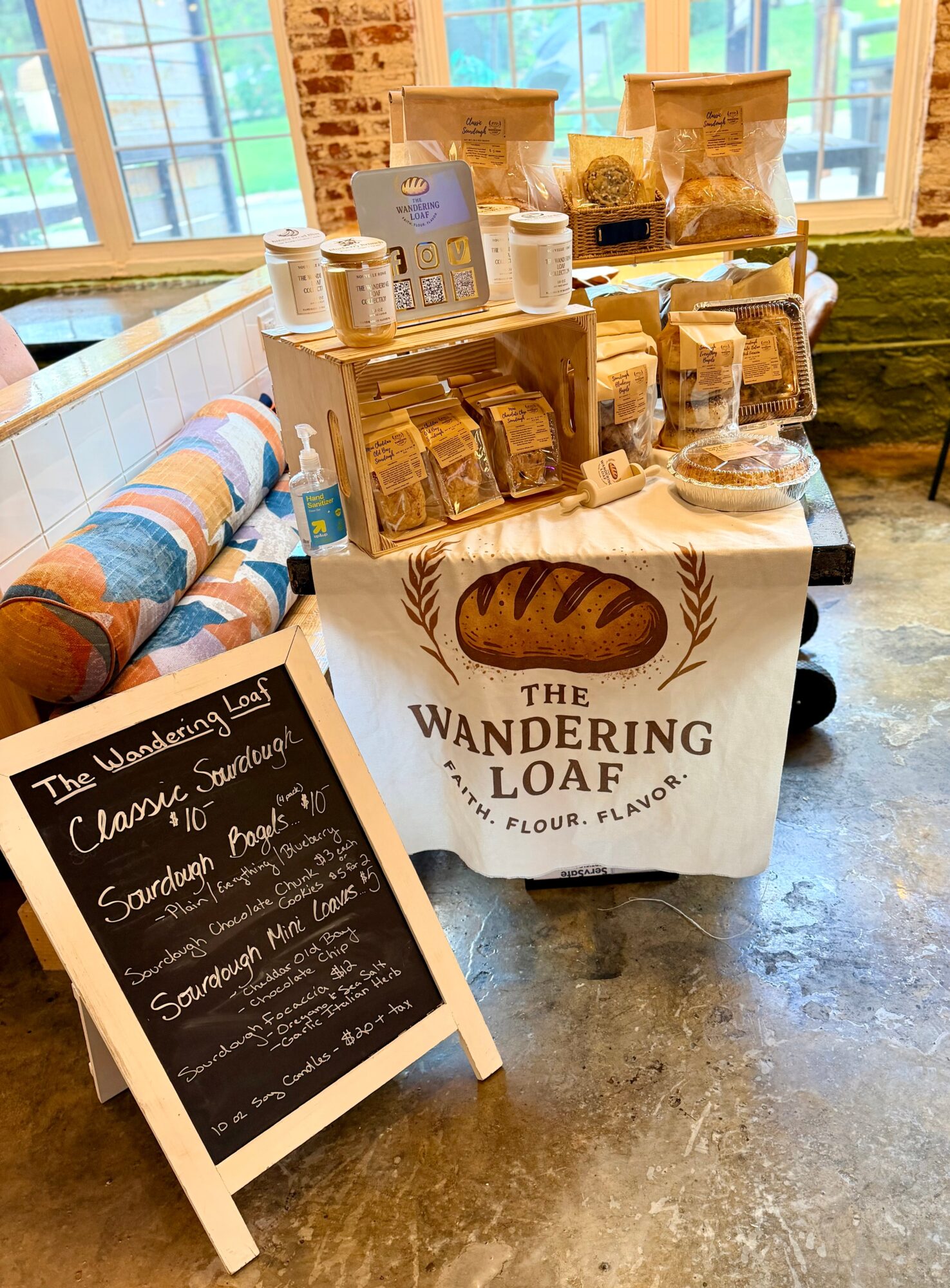 Display table with bread products and a sign for Wandering Loaf bakery, inside a room with large windows and brick wall.