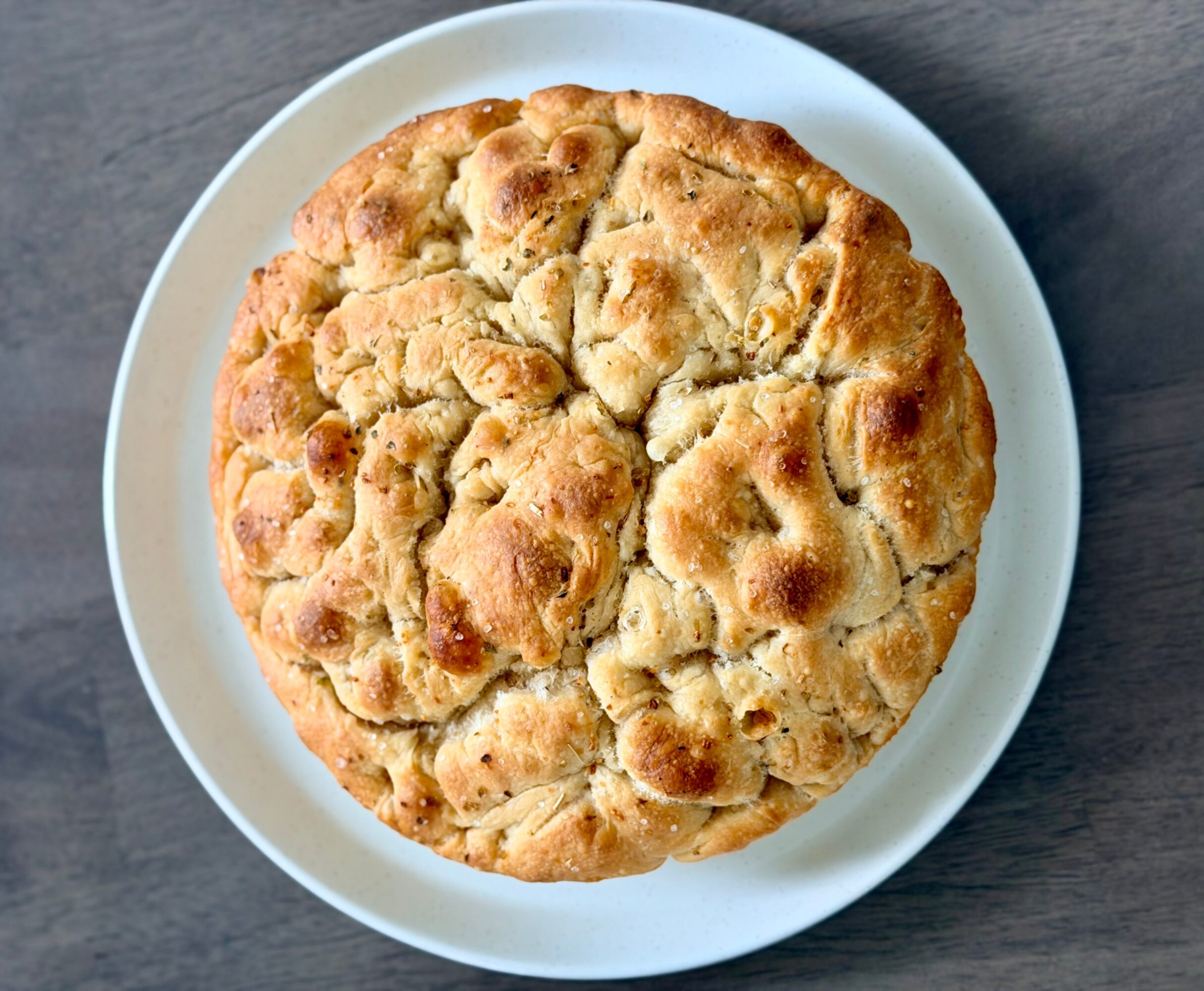 Round baked bread with a golden-brown crust on a white plate, placed on a dark surface.