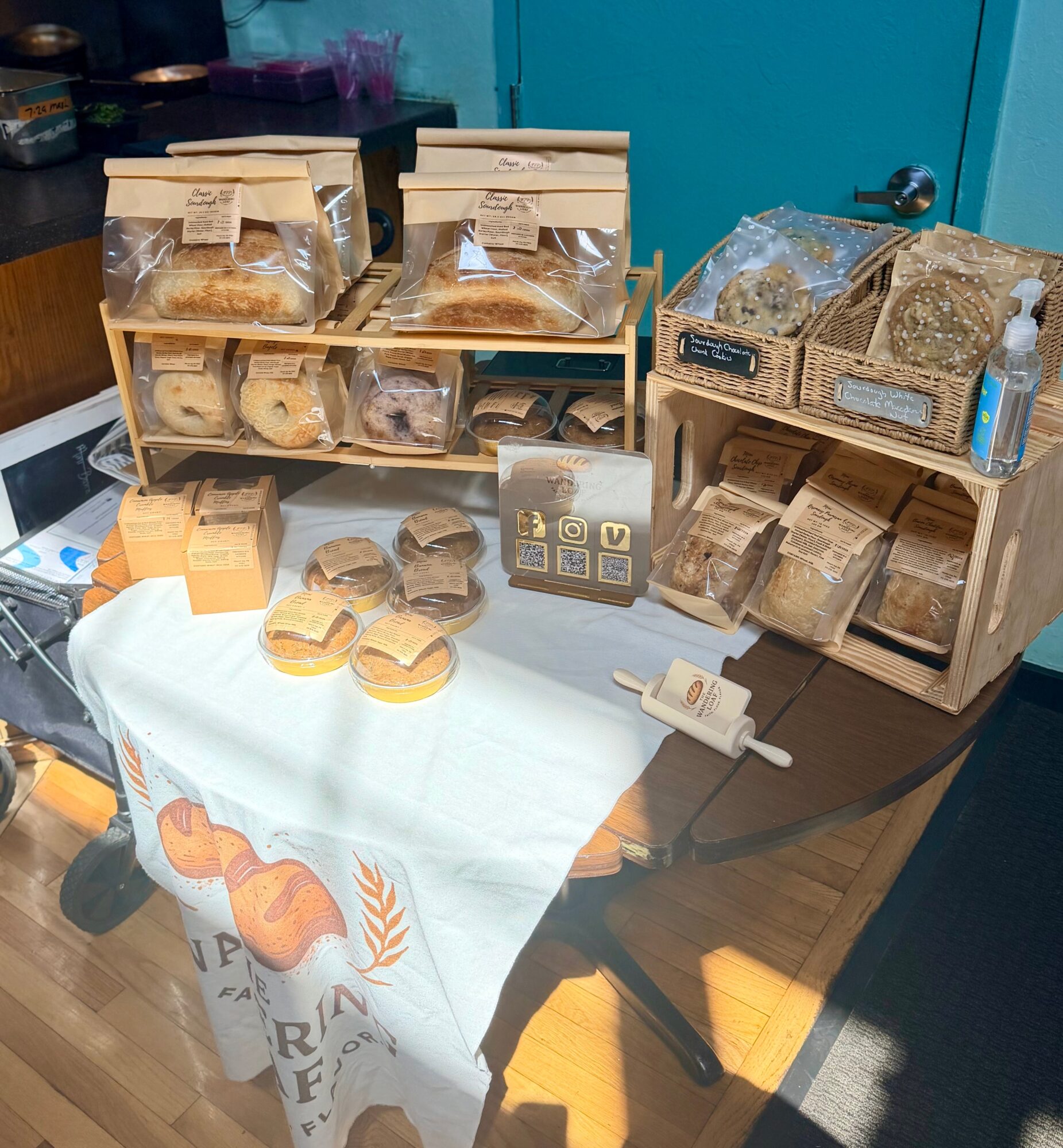 Display of packaged baked goods on a table with a cloth, with boxes and baskets holding bread and cookies, and a small sign.