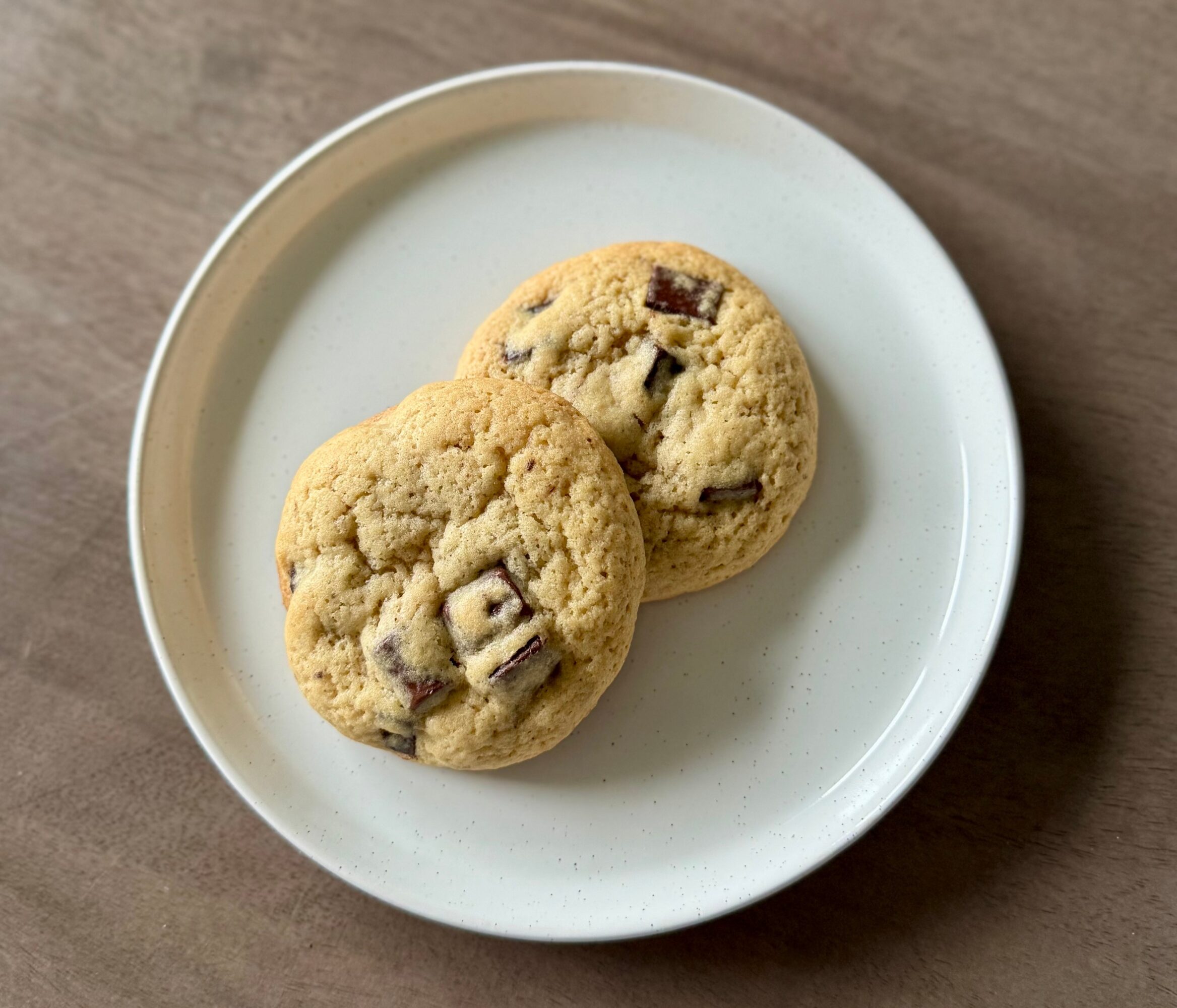 Two chocolate chip cookies on a white plate, placed on a wooden surface.