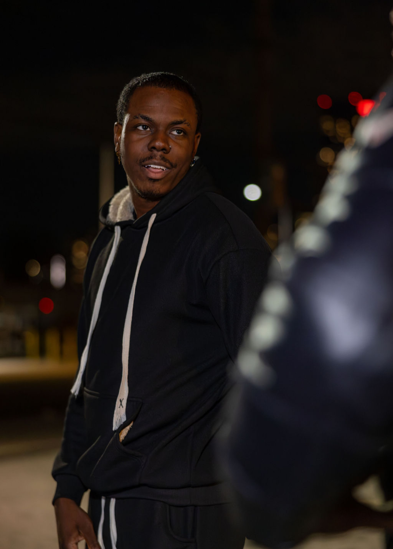 Young man with short hair and a beard, wearing a black hoodie, standing outdoors at night with city lights in background.
