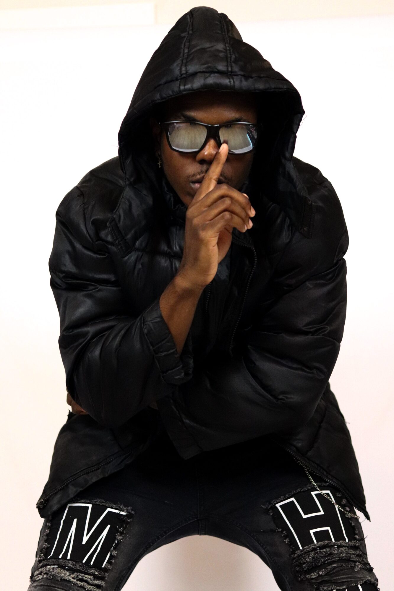Person wearing a black hoodie, sunglasses, and a black jacket, holding a finger to lips in a shushing gesture.