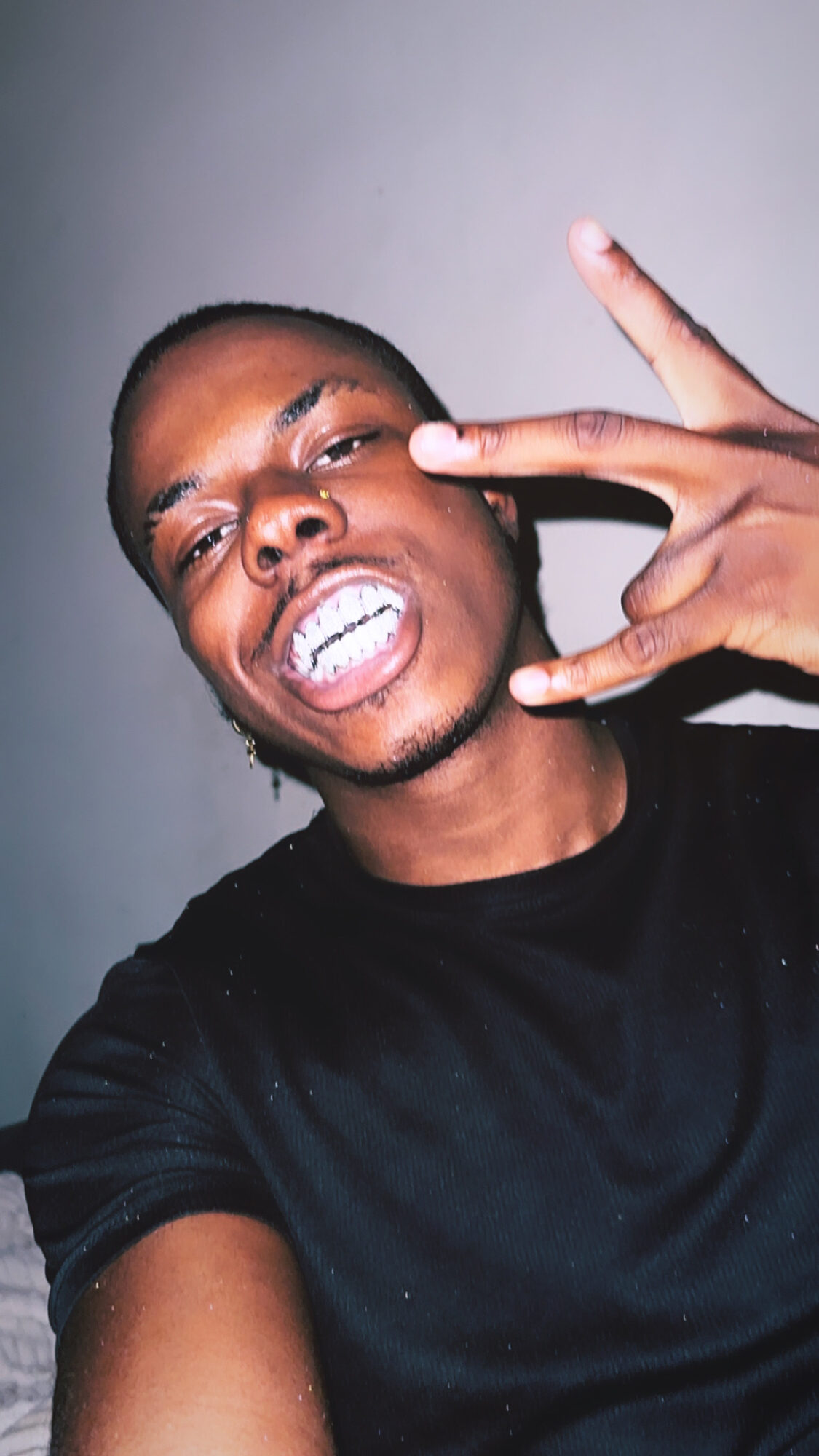 Young man with short hair making a peace sign near his face, smiling with teeth showing, wearing a black shirt.