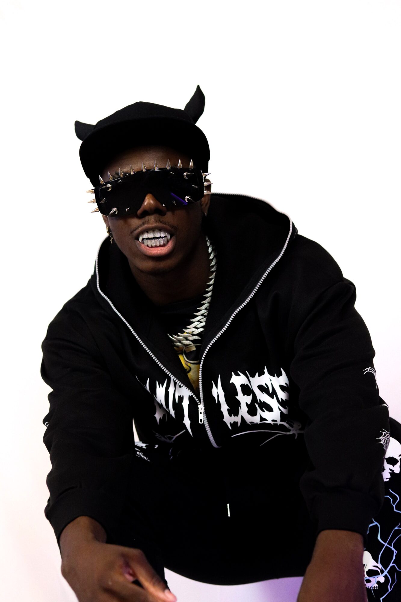 Person wearing sunglasses, a cap with ears, and a black hoodie with white text, smiling with visible teeth.