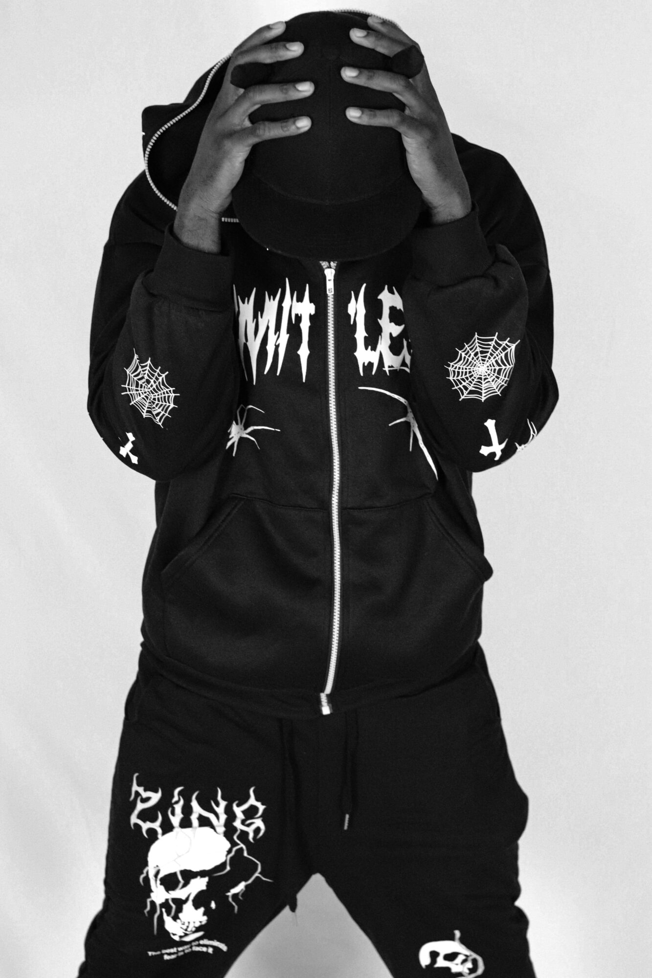 Person wearing a black hoodie with white graphics, holding their head, face obscured, standing against a plain background.